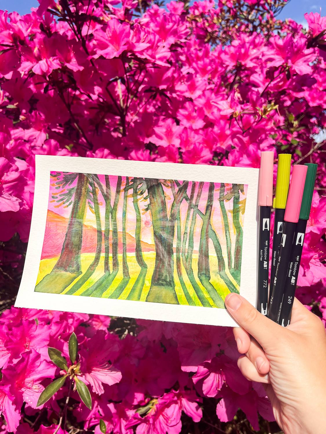Hand holding pink and green @tombowusa Dual Brush Pens, alongside finished artwork of a spring forest landscape painting in front of a blooming azalea bush