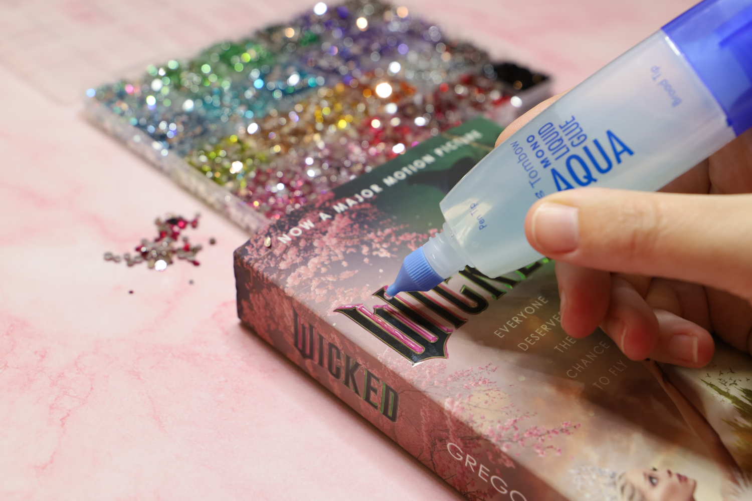 Hand holding @tombowusa MONO Aqua Liquid Glue, applying glue to a copy of a Wicked book to make a bedazzled book