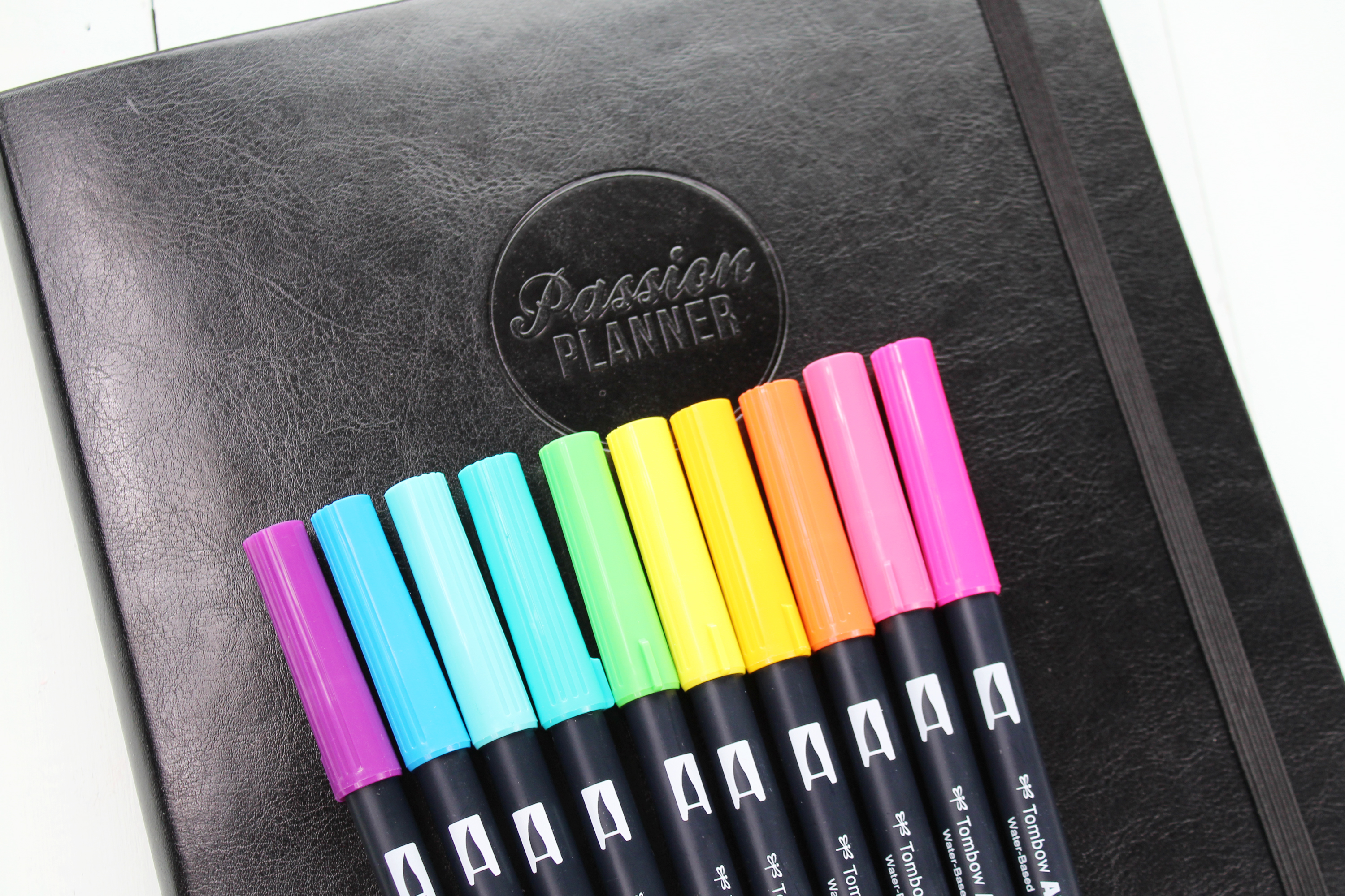 Tombow Dual Brush Pens in bright rainbow colors resting on a black leather Passion Planner showing logo.