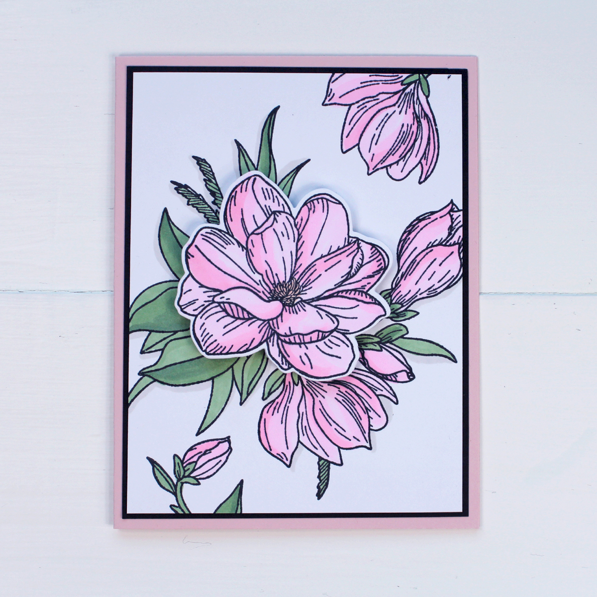 Blush pink magnolia flower card with green leaves on a white background. Mounted to a thin border of black cardstock and mounted to a blush pink folded card. 